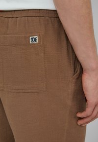 Pantalon tissé marron avec une taille élastique, présentant une poche latérale et une étiquette. La texture semble douce et légèrement surélevée.