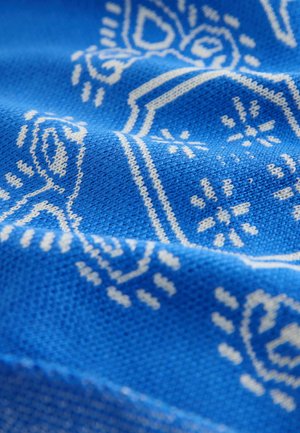 Mėlynos spalvos megzta medžiaga su balta sudėtinga geometrinių ir gėlių motyvų raštu, demonstruojanti tekstūruotą paviršių ir šešėliavimą.