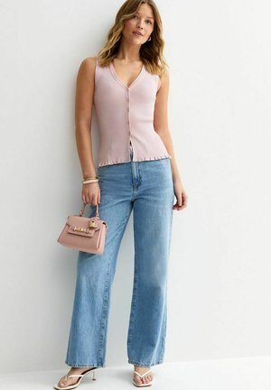 Femme portant un haut sans manches rose à boutons, un jean à jambes larges bleu clair, des sandales blanches à talons, et tenant un petit sac à main rose.