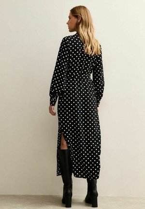 Robe longue noire à pois avec manches longues, présentant des fentes latérales. Tissu doux à la texture lisse. Portée avec des bottes noires montant jusqu'aux genoux.