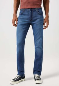 Ciemnoniebieskie dżinsy z prostymi nogawkami, z przednimi kieszeniami, kontrastowym przeszyciem i zużytym wykończeniem. W zestawie z czarnymi sneakersami.