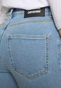 Vaqueros de mezclilla azul claro con una forma ajustada, con costuras prominentes, un bolsillo trasero y una etiqueta negra de marca en la cintura.