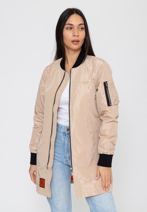 Frau trägt beige lange Bomberjacke mit schwarzen Bündchen und Reißverschluss, weißes Oberteil und hellblaue Jeans, blickt leicht zur Seite.