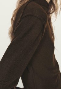 Primo piano di una persona che indossa un maglione lavorato a maglia marrone scuro con trama, con capelli lunghi e ondulati parzialmente visibili su uno sfondo neutro.