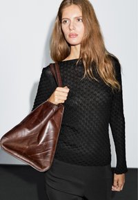 Black textured sweater with long sleeves, paired with a brown leather triangular bag. Model stands against a plain background.