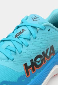 Ljusblå löparsko med texturerat ovanmaterial, svart "HOKA"-logotyp, orange accent och unik snörningsdesign; har andningsbart nätmaterial och vadderad krage.