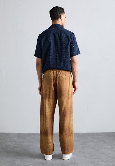 Navy blue short-sleeve shirt with textured pattern and cut-out details, paired with loose brown plaid trousers and white sneakers.