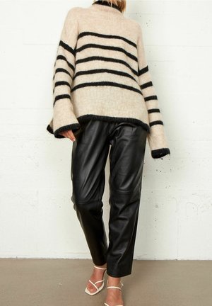 Persoon draagt een beige en zwart gestreepte trui, zwarte leren broek en witte sandalen met bandjes, staand tegen een witte muur.