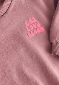 Ružová mikina s jemnou textúrou, ktorá má na prednej strane vystúpené viacfarebné písmená s nápisom „nech láska rozkvitne“. Rebrované manžety.