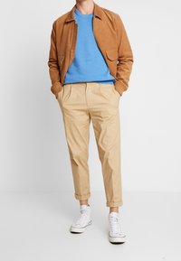 Hellbraune Cordjacke, blauer Strickpullover, beigefarbene Hose mit umgeschlagenen Bündchen und weiße Turnschuhe. Einfaches Design mit glatten Texturen.