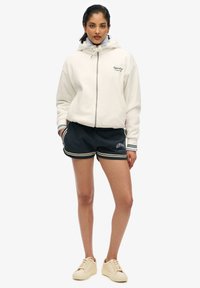 Witte zip-up hoodie met een trekkoord capuchon, gecombineerd met donkere shorts met beige strepen. Beige schoenen maken de outfit compleet.