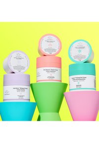 Quatre contenants de crème pour la peau dans des couleurs pastel empilés sur des formes géométriques vertes et bleues contre un fond jaune vif.