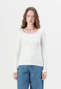 Hvit langermet topp med ribbestruktur, rund hals, og fem knapper foran. Kombinert med høytlivs blå denimjeans.