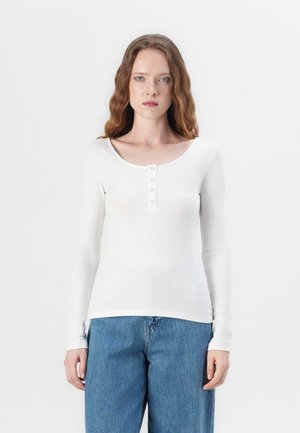 Hvit langermet topp med ribbestruktur, rund hals, og fem knapper foran. Kombinert med høytlivs blå denimjeans.