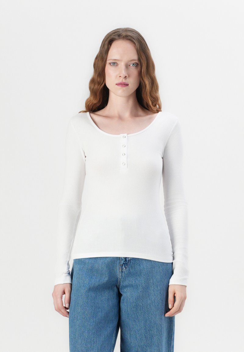 Hvit langermet topp med ribbestruktur, rund hals, og fem knapper foran. Kombinert med høytlivs blå denimjeans.