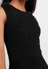 Robe noire en tricot sans manches avec des motifs texturés, présentant des designs géométriques en relief et une finition côtelée en haut et en bas.