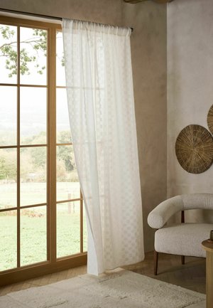 Tenda bianca trasparente parzialmente tirata su una finestra con cornice in legno che si affaccia su un paesaggio verde, con una poltrona curva beige e decorazioni da parete intrecciate nelle vicinanze.
