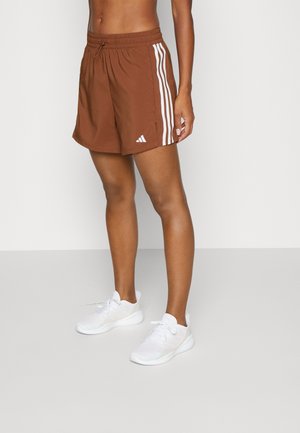 Persoon draagt bruine sportshorts met witte strepen en witte sneakers, staand tegen een effen lichte achtergrond.