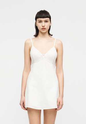 Joven con cabello corto y oscuro que lleva un minivestido blanco sin mangas con tirantes finos, posando frente a un fondo blanco liso.