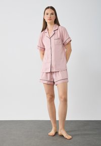 Roze satijnen pyjama set met een knoopsluiting aan de voorkant, korte mouwen en een borstzak, gecombineerd met bijpassende shortjes afgewerkt met zwarte bies.