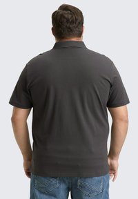 Mężczyzna ubrany w ciemnoszary krótki polo i niebieskie dżinsy, stojący plecami do kamery na jednolitym tle.