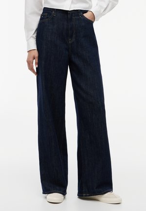 Osoba nosiaca tmavomodré široké rifle s prednými vreckami a bielymi teniskami, stojaca s jednou rukou vo vrecku.