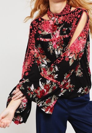 Sort bluse med lange ærmer med rød blomsterbroderi, udskårne skulderdetaljer og flagrende ærmer, kombineret med mørkeblå bukser.