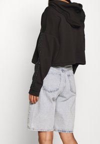 Felpa nera corta con cappuccio, ornata da applicazioni di strass sulle maniche. Abbinata a pantaloncini di jeans azzurri, che mostrano una vestibilità casual.