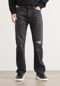 Jean en denim noir avec une coupe décontractée, présentant un détail déchiré au genou droit et un design classique à cinq poches. Associé à des baskets noires.