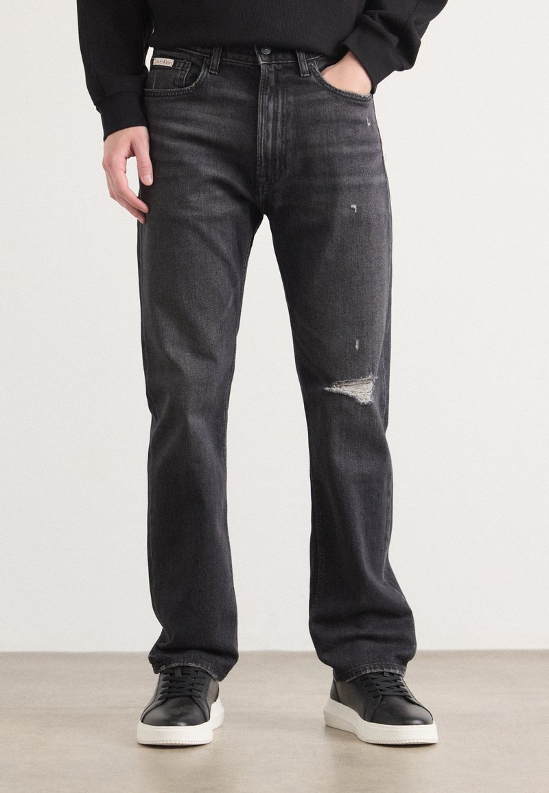 Jean en denim noir avec une coupe décontractée, présentant un détail déchiré au genou droit et un design classique à cinq poches. Associé à des baskets noires.
