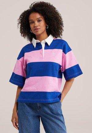 Mujer con cabello rizado que lleva una camiseta de rugby a rayas azules y rosas con cuello blanco y vaqueros azules, de pie con una mano en el bolsillo.