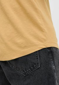 Kortærmet, sennepsgul t-shirt med en let struktureret stof. Parret med mørkegrå denim jeans med en baglomme.