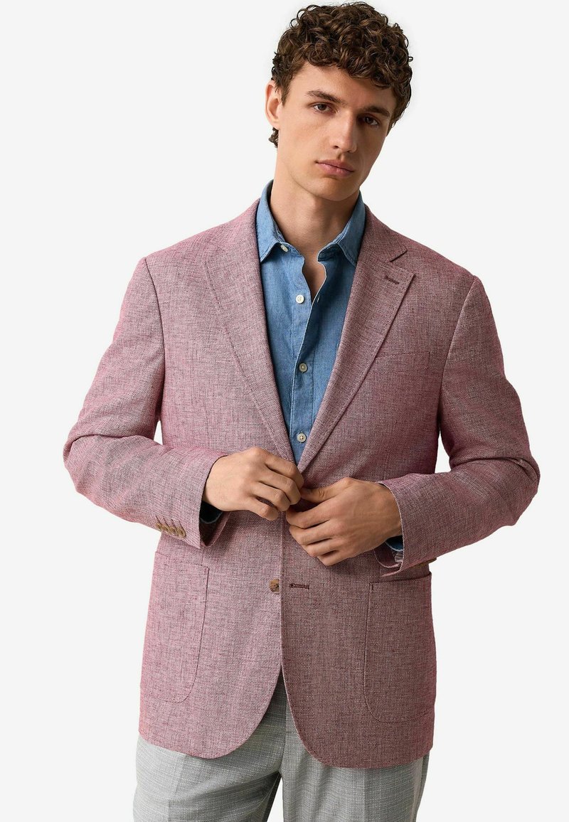 Blazer texturé rouge avec deux poches avant, fermeture à bouton unique et revers, porté sur une chemise en denim bleue.