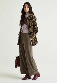 Giacca lunga a stampa mimetica con pantaloni verde oliva, camicia a righe e stivaletti alla caviglia in bordeaux testurizzato, abbinati a una borsa bordeaux.