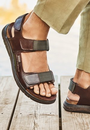 Persona con sandalias de cuero marrón con correas oscuras ajustables, de pie sobre una superficie de madera cerca del agua, vestida con pantalones verde claro.