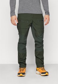 Zielone spodnie outdoorowe z funkcjonalnymi, zapinanymi na zamek kieszeniami, łączące trwały materiał z elastycznym krojem, w zestawie z pomarańczowymi butami trekkingowymi.