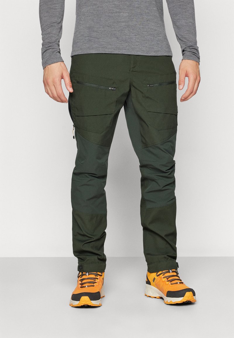Zielone spodnie outdoorowe z funkcjonalnymi, zapinanymi na zamek kieszeniami, łączące trwały materiał z elastycznym krojem, w zestawie z pomarańczowymi butami trekkingowymi.