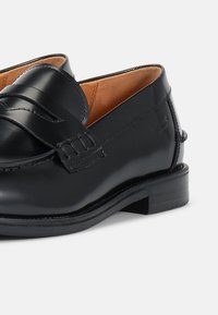 Μαύρο δερμάτινο λουστρίνι με λεία επιφάνεια, με στρογγυλή μύτη, ζώνη σέλας και αντίθετο καφέ επένδυση. Χαμηλό μπλοκ τακούνι.