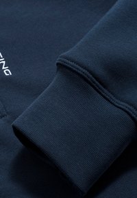 Sudadera de color azul marino con textura de canalé y puños. Presenta un bordado de logo blanco en la parte frontal. Vista en primer plano que resalta los detalles.