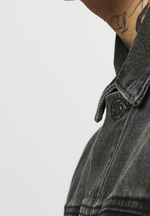 Nærbillede af en persons nakke og skulder iført en mørk denimjakke med en sort knap, der viser en delvis tatovering på nakken.