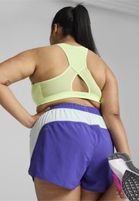 Sutien sport light green cu design încrucișat pe spate, asociat cu pantaloni scurți mov și adidași gri. Pantalonii scurți au o bandă de talie contrastantă de culoare albastru deschis.