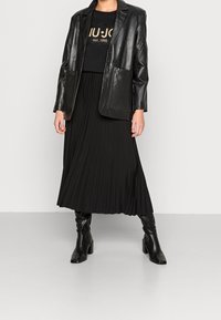 Blazer de cuero negro sobre una camiseta negra con texto dorado, combinado con una falda negra con pliegues y botas de tacón alto negras hasta la rodilla.