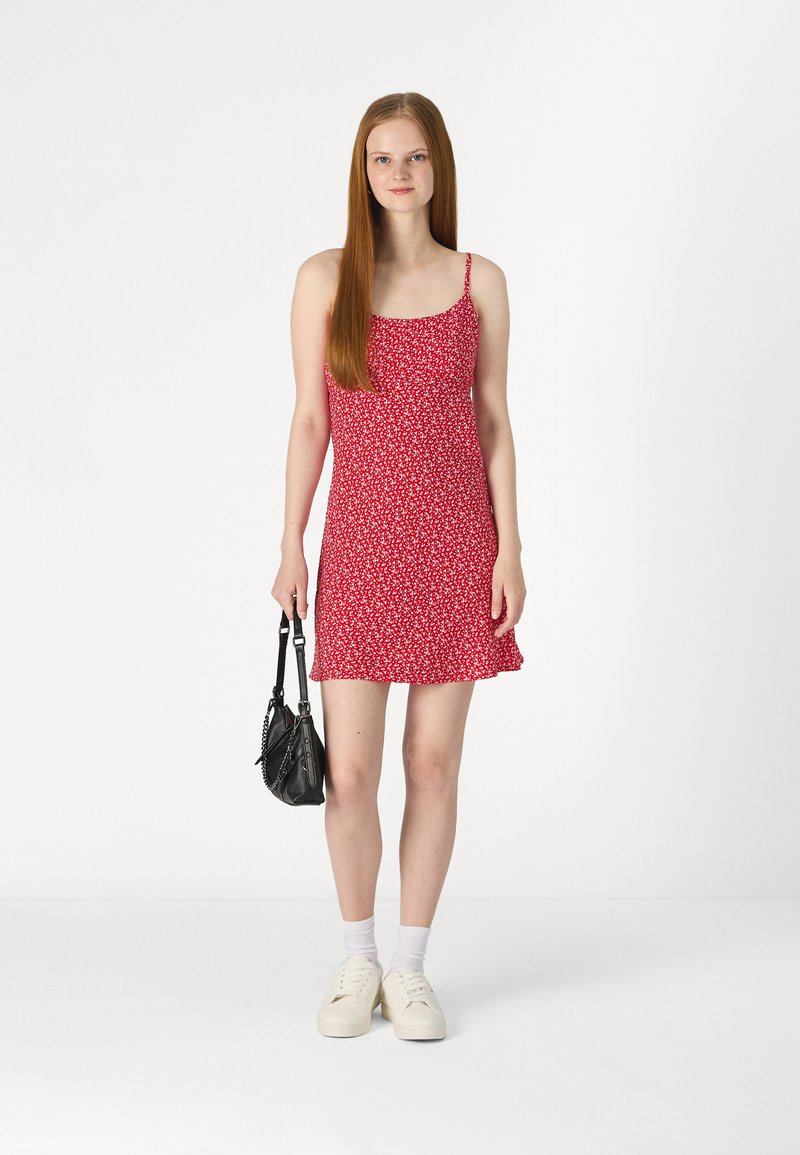 Rotes Kleid mit floralem Muster, dünnen Trägern, tailliertem Oberteil und ausgestelltem Rock. Trägt eine schwarze Handtasche und leichte Sneakers.