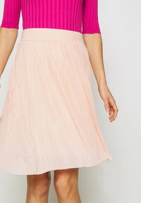 Jasnoróżowa plisowana spódnica z gładkim pasem, o lejącym się materiale tworzącym delikatny ruch. Częściowo widoczny prążkowany różowy top.