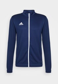 Marineblauwe sportjack met hoge kraag, volledige rits aan de voorkant en witte accenten. Beschikt over het klassieke drie-strepen adidas-logo op de borst.