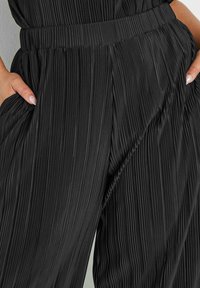 Svart plisserade byxor med bred midja, med vertikala veck för textur och sidofickor integrerade i designen.