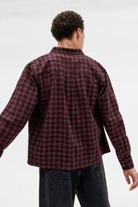 Pessoa com cabelo curto e cacheado, vestindo uma camisa xadrez vermelha e preta e jeans pretos, vista de costas contra um fundo branco liso.
