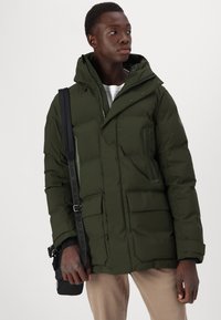 Dunkelgrüne Steppjacke mit Reißverschluss und Druckknopfverschluss, verstellbarem Kapuze, Seitentaschen und strukturierter Oberfläche, getragen mit einer schwarzen Umhängetasche.