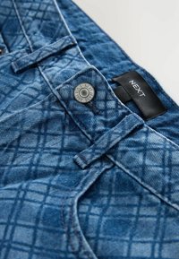 Jeans din denim albastru cu un model matlasat, având un închizător cu buton ridicat și un eticheta neagră cu inscripția "NEXT". Țesătura are un aspect texturat.