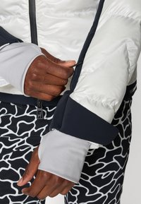 Chaqueta blanca acolchada con detalles en azul oscuro y mangas con orificios para el pulgar, llevada sobre pantalones a rayas en blanco y negro. Detalle de cremallera en la chaqueta.